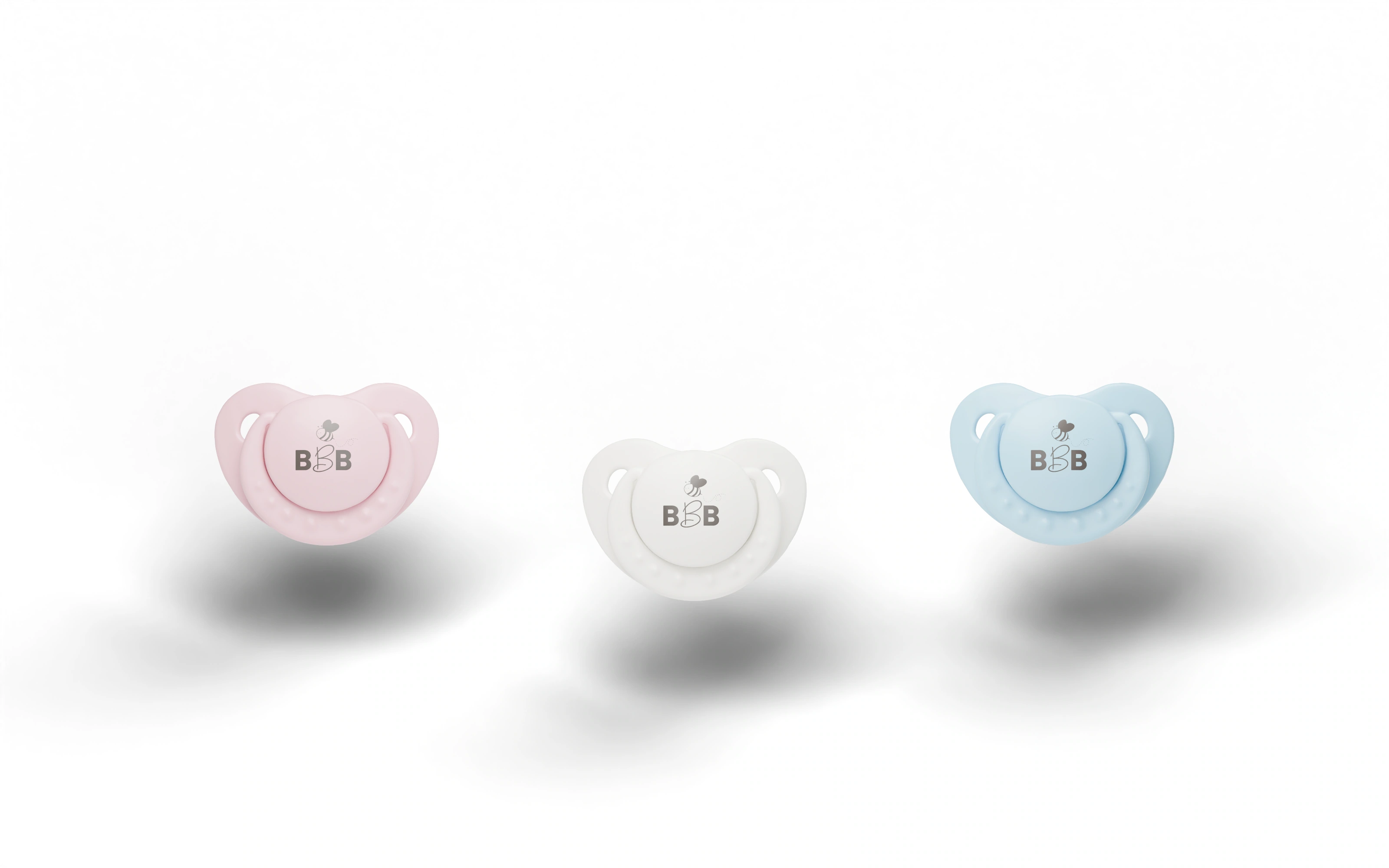 Three Baby Bee Blossom pacifiers hovering above soft shadows, logos facing forward: pink on the left, white in the middle, and blue on the right.